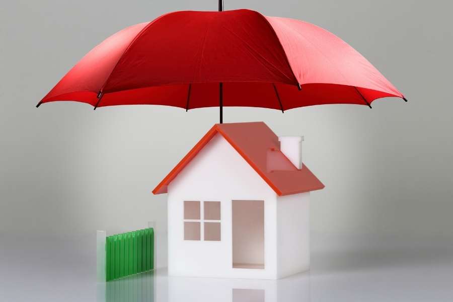 Une petite maquette de maison blanche avec un toit rouge est protégée par un grand parapluie rouge ouvert.