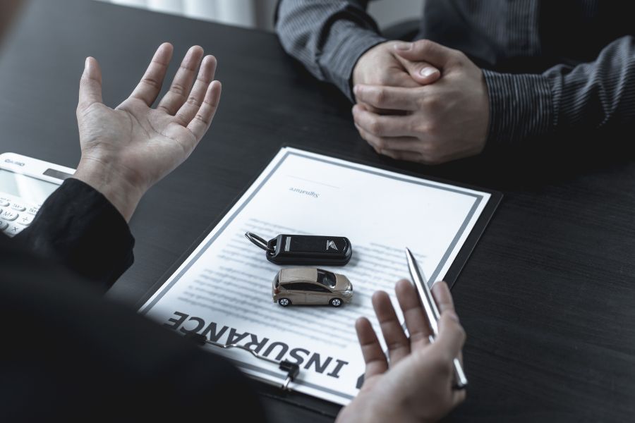 Un agent d'assurance et un client discutent autour d'un bureau sombre, avec un contrat d'assurance auto, une miniature de voiture et des clés visibles.